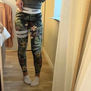 Mönstrade leggings från Stronger - Snygga och bekväma leggings från Stronger med tropiskt blommönster i grönt, rosa och vitt. Midjan är en elastisk linning med märkeslogga. Perfekta för träning eller en avslappnad dag hemma. De är i mycket bra skick och passar perfekt för vilken årstid som helst!