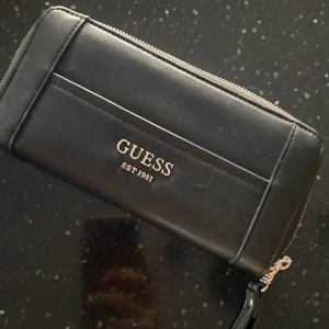 Svart plånbok från Guess - Snygg och stilren svart plånbok från Guess med dragkedja. Perfekt för att hålla ordning på kort och kontanter. Märket är tryckt i guld på framsidan. Plånboken har flera fack och en praktisk design.