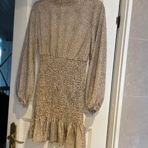 Beige prickig klänning - Säljer en superfin beige halvpolo klänning med små prickar. Den har långa ärmar och en smickrande tight passform med volangdetalj längst ner. Perfekt för både vardag och fest! 🥰