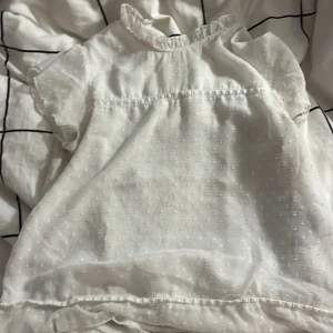 Vit blus med volangdetaljer - Söt vit blus med korta ärmar och volangdetaljer vid halsen. Blusen har ett diskret prickigt mönster och är perfekt för vår och sommar. Den är luftig och bekväm, idealisk för både vardag och festliga tillfällen.