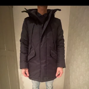 Mörkblå parkajacka från Krakatau nypris 4500kr  - Säljer en mörkblå parkajacka från Krakatau i mycket bra skick. Jackan är perfekt för vintern med sin långa passform och quiltade insida. Den har en praktisk dragkedja och knappar framtill samt en huva för extra skydd mot kyla. Reflekterande detaljer på baksidan ger en cool touch. Perfekt för kalla dagar! ❄️nypris ligger runt 4500kr 