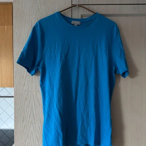 Blå t-shirt från COS - Storlek S - Säljer en snygg blå t-shirt från COS i storlek S. Den är gjord av mjuk bomull och har en klassisk rund halsringning. Perfekt för både vardag och avslappnade tillfällen. T-shirten är i mycket bra skick och redo för en ny ägare!
