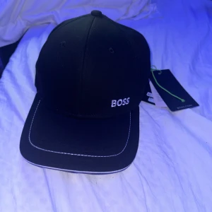 BOSS keps - Säljer en stilren svart keps från BOSS helt ny aldrig använd i bomull. Kepsen har en broderad BOSS-logga framtill och justerbart spänne bak. Perfekt för både vardag och sportiga aktiviteter. Helt ny med taggar kvar!