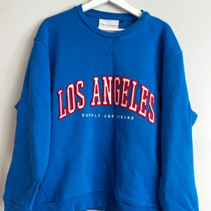 Blå sweatshirt från Supply & Demand - Säljer en snygg blå sweatshirt från Supply & Demand med texten 'Los Angeles' i rött och vitt på framsidan. Tröjan är i storlek S och har en oversized passform. Mycket bra skick förutom en defekt på tröjan (se bild)