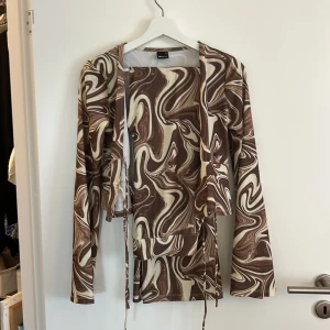 Brunmönstrat set från Gina Tricot - Säljer en supercool brunmönstrad omlottjacka med tillhörande byxor från Gina Tricot. Den har ett unikt swirlmönster i brunt och beige, perfekt för att sticka ut. Knytblusen är långärmad och har knytband framtill för en snygg passform. Perfekt för höst och vår!