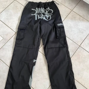 Svarta cargobyxor med tryck - Säljer ett par svarta cargobyxor med coolt tryck i grått från minus two. Byxorna har flera fickor och snörning i midjan för en bekväm passform. Perfekta för en avslappnad och stilren look. skriv vid frågor💗