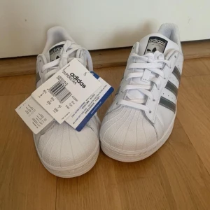 Adidas Superstar  - Säljer ett par Adidas Superstar sneakers i vitt med silvriga ränder. De är helt nya och har fortfarande taggarna kvar💗