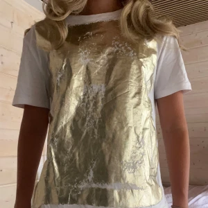 Vit t-shirt med guldtryck - Säljer en cool vit t-shirt med ett stort guldtryck på framsidan. Den är kortärmad och perfekt för en avslappnad men ändå stilren look. Passar bra till både jeans och shorts. Perfekt för sommaren eller en festkväll! ✨