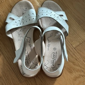 Vita komfortabla sandaler i storlek 38 - 100 kr - Beskrivning: Säljer ett par vita sandaler i storlek 38. De har breda remmar med dekorativa utskärningar och en låg kilklack för extra komfort. Perfekta för sommarens alla tillfällen, både vardag
