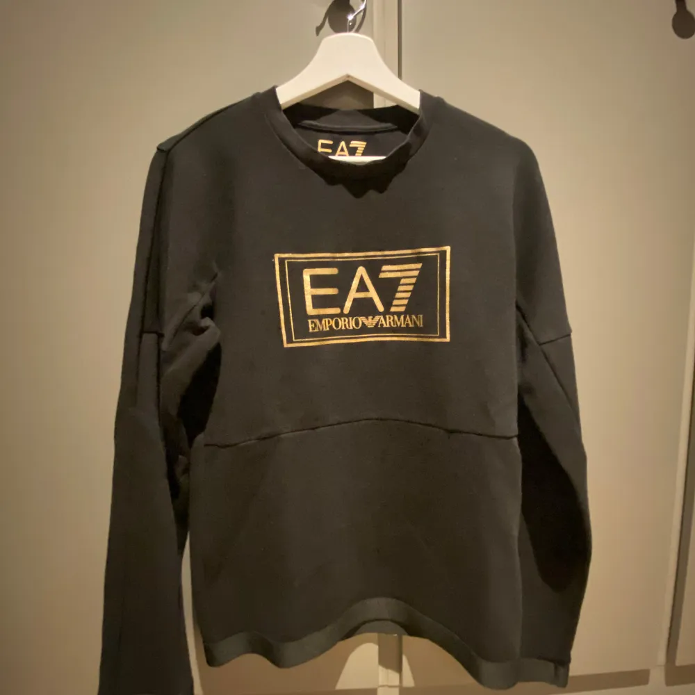 Snygg svart tröja från Emporio Armani med guldtryck av EA7-loggan på bröstet. Tröjan har en klassisk rund hals och långa ärmar, perfekt för en stilren look. Passar bra till både jeans och chinos.. Neuletakit & Villapaidat.