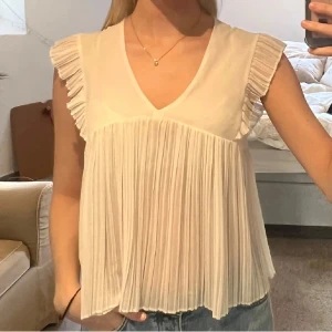 Zara Vit blus med volangdetaljer - Säljer en söt vit blus från Zara med volangdetaljer på ärmarna. Blusen har en v-ringad hals och är ärmlös, vilket gör den perfekt för varmare dagar. Passar bra till både jeans och kjol. Knappt använd! 