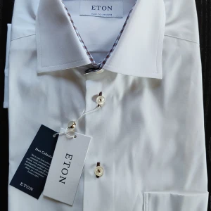 Vit skjorta från Eton - Elegant vit skjorta från Eton med klassisk design och stilrena knappar. Perfekt för en sofistikerad look. Tillverkad med svensk hantverkstradition. Helt ny! Mått:Total length: 84 cm Width from armpit to armpit: 69 cm Waist width: 68 cm Collar width: 51 cm Sleeve length from armpit: 53 cm