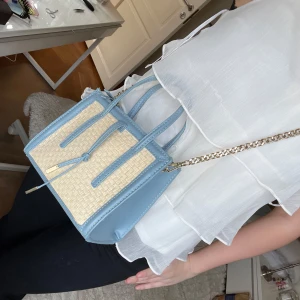 Blå och beige axelväska med kedja - Snygg axelväska i blått och beige med flätad design. Väskan har en elegant kedjerem och dragkedja upptill. Perfekt för att ge din outfit en stilfull touch.