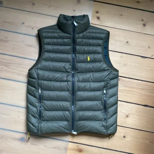 Grön dunväst från Polo Ralph Lauren - Säljer en snygg grön dunväst från Polo Ralph Lauren. Västen har en dragkedja framtill och en liten gul broderad logga på bröstet. Perfekt för kyligare dagar och ger en stilren look.