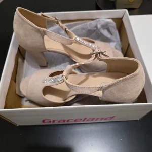 Beige klackskor från Graceland - Eleganta beige klackskor från Graceland med öppen tå och T-rem dekorerad med glittrande stenar. Skorna har en bekväm klack och justerbart spänne runt ankeln. Helt oanvända 