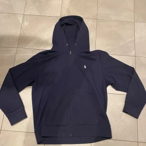 Mörkblå hoodie från Polo Ralph Lauren - Säljer en snygg mörkblå hoodie från Polo Ralph Lauren med dragkedja och det klassiska broderade logotypen på bröstet. Perfekt för en avslappnad stil. Den har långa ärmar och en praktisk huva.