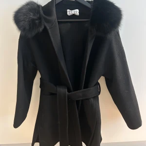 Svart kappa från fur in Stockholm  - Elegant svart kappa från fur in Stockholm med lyxiga pälsdetaljer på huvan. Kappan har ett bälte i midjan för en snygg passform och långa ärmar. Perfekt för kyligare dagar när du vill känna dig både varm och stilren Men funkar även på våren.  Endast testad men tyvärr inga lappar kvar. Super fint skick.  Äkta päls.
