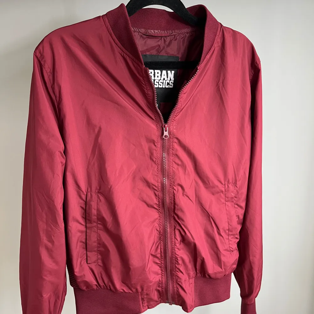 Snygg röd bomberjacka från Urban Classics med dragkedja framtill. Jackan har en klassisk bomberstil med ribbade muddar vid ärmslut och nederkant. Perfekt för en cool och avslappnad look.. Takit.