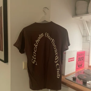 Brun t-shirt från Stockholm Surfboard Club - Snygg brun t-shirt från Stockholm Surfboard Club med tryck på baksidan. Perfekt för en avslappnad stil. T-shirten har en klassisk passform och rund hals.
