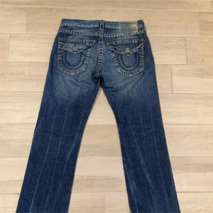 Blå jeans från True Religion - Säljer ett par snygga blå jeans från True Religion. De har en straight fit och är dekorerade med slitningar för en cool look. Jeansen har knappar och dragkedja samt de ikoniska bakfickorna med broderade detaljer.