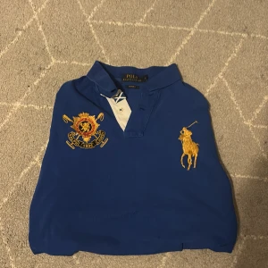 Blå pikétröja från Ralph Lauren - Säljer en snygg blå pikétröja från Ralph Lauren med broderad gul logotyp på bröstet och ett emblem på andra sidan. Tröjan har en klassisk krage och knappar framtill. Siffran 3 är broderad på ärmarna. Perfekt för en stilren look! Storlek är L i man storlek. Det är bara att fråga om ni har några mer frågor. Skick är 8/10 skulle ja väl säga.