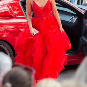 Röd balklänning med spetsdetaljer - Säljer min fantastiska röda balklänning med vacker spets och volanger. Klänningen har en djup v-ringning och är ärmlös, perfekt för balen. Den har en fyllig kjol med flera lager som ger en dramatisk look. 