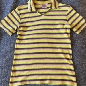 Randig pikétröja från INDISKA - Säljer en retroinspirerad randig pikétröja från INDISKA i gul, brun och beige. Tröjan har korta ärmar och en klassisk krage med v-ringning. Perfekt för en avslappnad stil med en touch av vintage.