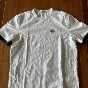 Moncler t shirt  - Säljer en stilren vit t-shirt från Moncler med korta ärmar och ett broderat Moncler-märke på bröstet. Perfekt för en avslappnad och trendig look. T-shirten har en klassisk passform och är tillverkad i mjukt material.