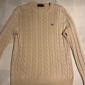 Beige kabelstickad tröja från Morris strl M - Säljer en stilren beige kabelstickad tröja från Morris i 100% bomull. Tröjan har en rund halsringning och ett diskret broderat emblem på bröstet. Perfekt för en klassisk och avslappnad look. Säljer pga för stor strl. 