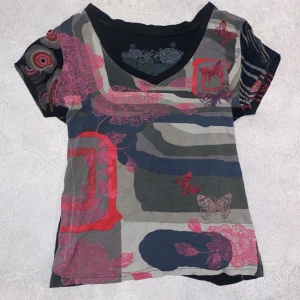 Mönstrad topp från Desigual - Säljer en unik och färgglad topp från Desigual med ett abstrakt mönster i rött, rosa och grönt. Toppen har en v-ringad hals och korta ärmar med olika mönster på fram- och baksidan. Perfekt för dig som gillar att sticka ut!