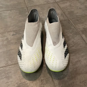 Vita fotbollsskor från Adidas - Snygga vita fotbollsskor från Adidas med svarta och neongröna detaljer. Skorna har en strömlinjeformad design utan snörning och en greppvänlig sula med dobbar för optimalt fäste på planen. Perfekta för den seriösa fotbollsspelaren.