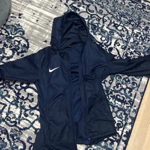 Mörkblå vindjacka från Nike - Säljer en mörkblå vindjacka från Nike med huva och dragkedja. Jackan har en vit Nike-logga på bröstet och är perfekt för blåsiga dagar. Den är långärmad och har en sportig look.