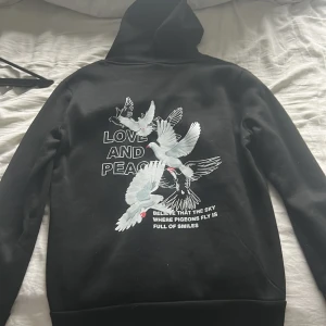 Svart hoodie med duvmotiv - Säljer en svart hoodie med ett stort tryck av duvor och texten 'LOVE AND PEACE' på ryggen. Den har en klassisk känguruficka framtill och en bekväm huva. Perfekt för en avslappnad stil.