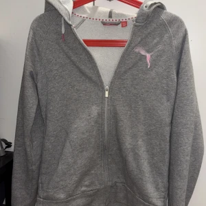 Grå hoodie från Puma - Säljer en grå hoodie från Puma med dragkedja och rosa logga på bröstet. Tröjan har en mysig huva och långa ärmar, perfekt för kyliga dagar. Den är tillverkad i ett mjukt material som känns skönt mot huden. Passar bra till både träning och vardag. SÄLJER DEN PGA ATT DEN ÄR FÖR STOR OCH ANVÄNDS INTE LÄNGRE