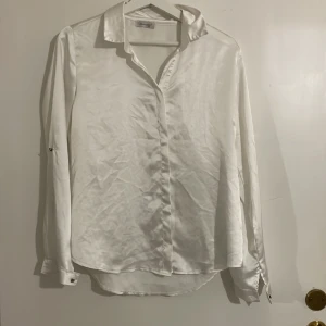Vit sidenblus - Elegant vit blus i siden med klassisk krage och långa ärmar. Perfekt för en stilren look. Blusen har en mjuk och glansig yta som ger en lyxig känsla.