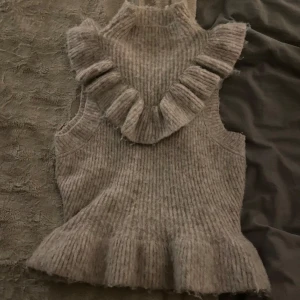 Beige stickad topp med volang - Säljer en mysig beige stickad topp med volangdetaljer vid axlarna och nederkanten. Toppen är ärmlös och har en hög krage.  Passar både till jeans och kjol för en stilren look. Köpt från zara.