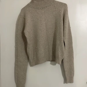 Beige stickad polotröja med pärlor - Säljer en elegant beige stickad polotröja dekorerad med pärlor. Tröjan har långa ärmar och en ribbad polokrage som ger en stilren look. Perfekt för kyligare dagar och ger en touch av lyx till din outfit. Står XL/L på tröjan men skulle säga M/S