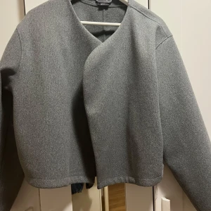 Grå jacka från Gina Tricot - Snygg grå jacka från Gina Tricot i storlek M. Jackan har en stilren design med en omlott-look och långa ärmar. Perfekt för en chic och minimalistisk stil.