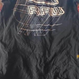 Svart t-shirt från G-Star RAW - Snygg svart t-shirt från G-Star RAW med ett coolt grafiskt tryck på framsidan. Tillverkad av hållbara material, perfekt för en stilren look. Märkesdetaljer på sidan ger en extra touch. Passar perfekt för dig som gillar streetwear.