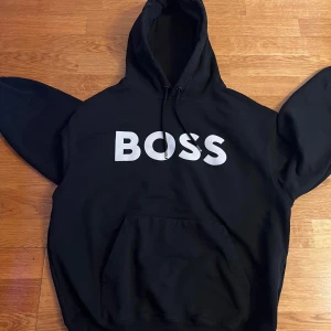 Svart hoodie från Boss - Snygg svart hoodie från Boss med stor vit logga på framsidan. Den har en klassisk design med huva och känguruficka. Perfekt för en avslappnad stil.