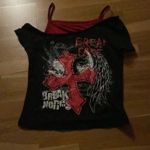 Svart offshoulder topp med tryck - Säljer en svart offshoulder topp med ett stort rött kors och texten 'Break Free' i vitt och rött. Toppen har smala axelband och en cool, edgy design. Perfekt för en unik stil! Bara använd en gång och det var när jag provade den