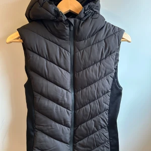 Svart quiltad väst från Zara - Snygg svart quiltad väst från Zara med dragkedja och huva. Västen har en stilren design med diagonala sömmar.