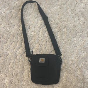 Svart axelväska från Carhartt - Säljer en stilren svart axelväska från Carhartt. Väskan har en justerbar axelrem och en praktisk ficka med dragkedja framtill. Perfekt för att bära med sig det nödvändigaste på ett smidigt sätt.