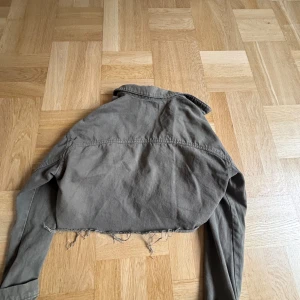 Grå croppad jacka med fransar - Säljer en grå croppad jacka från Zara med fransiga kanter. Jackan har långa ärmar och två bröstfickor med knappar. Perfekt för en avslappnad stil.