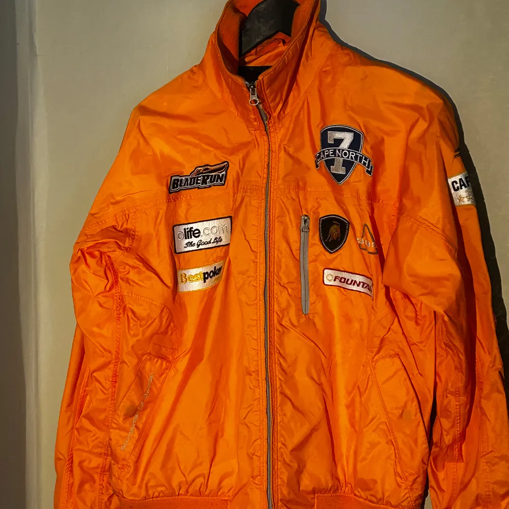 orange racingjacka från SOS med flera patches och broderier. Dragkedja och är långärmad, perfekt för dig som gillar en sportig stil. Bästa vodka märket 'Capenorth' finns även😉. Takit.