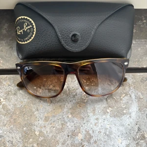 Ray-Ban Boyfriend Solglasögon - Snygga solglasögon från Ray-Ban med en klassisk brun sköldpaddsmönstrad båge. De har en stilren design med gradalins som går från mörkt till ljust. Perfekta för soliga dagar och kommer med ett svart fodral för säker förvaring. Även putsduk ingår. Sparsamt använda och i princip nyskick. Meddela vid behov av fler bilder. Pris kan diskuteras men endast rimliga bud. 