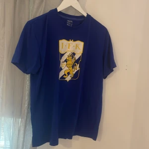 IFK tröja  - Säljer en blå t-shirt från landslaget IFK med en stor IFK-logga på framsidan. T-shirten är kortärmad och tillverkad i ett mjukt material. Perfekt för sportiga tillfällen eller för att visa ditt stöd för laget.