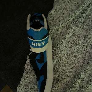 Nike sneakers i blått och svart - Snygga Nike sneakers i en fräsch kombination av blått och svart. Skorna har en klassisk design med vit sula och tydlig Nike-logga. Perfekta för en sportig look.