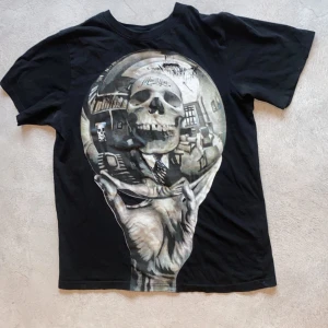 Svart t-shirt med dödskallemotiv - Cool svart t-shirt med ett stort, detaljerat dödskallemotiv på framsidan. Motivet har en konstnärlig stil med en hand som håller en sfär där dödskallen syns. Perfekt för dig som gillar unika och iögonfallande tryck.