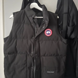  Canada Goose väst S - Säljer en svart dunväst från Canada Goose med broderad logotyp på bröstet. Västen har både dragkedja och knappar framtill för extra skydd mot kyla. Perfekt för kyliga dagar.INGA SKADOR 